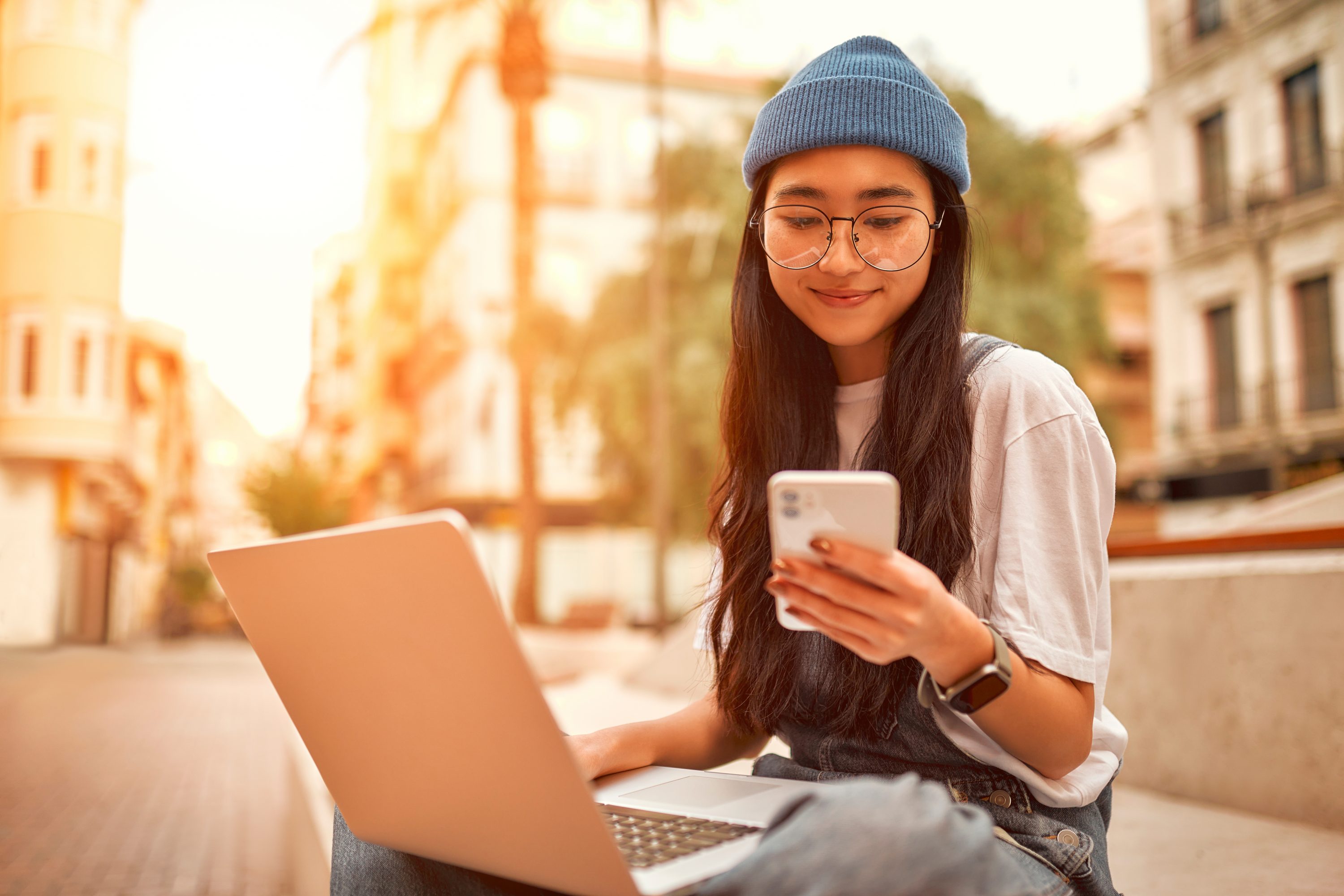 Portrait of asian tourist female hipster student in white shirt sitting on bench with laptop talking on the phone. Female student studying near campus. Freelancer working on the street.