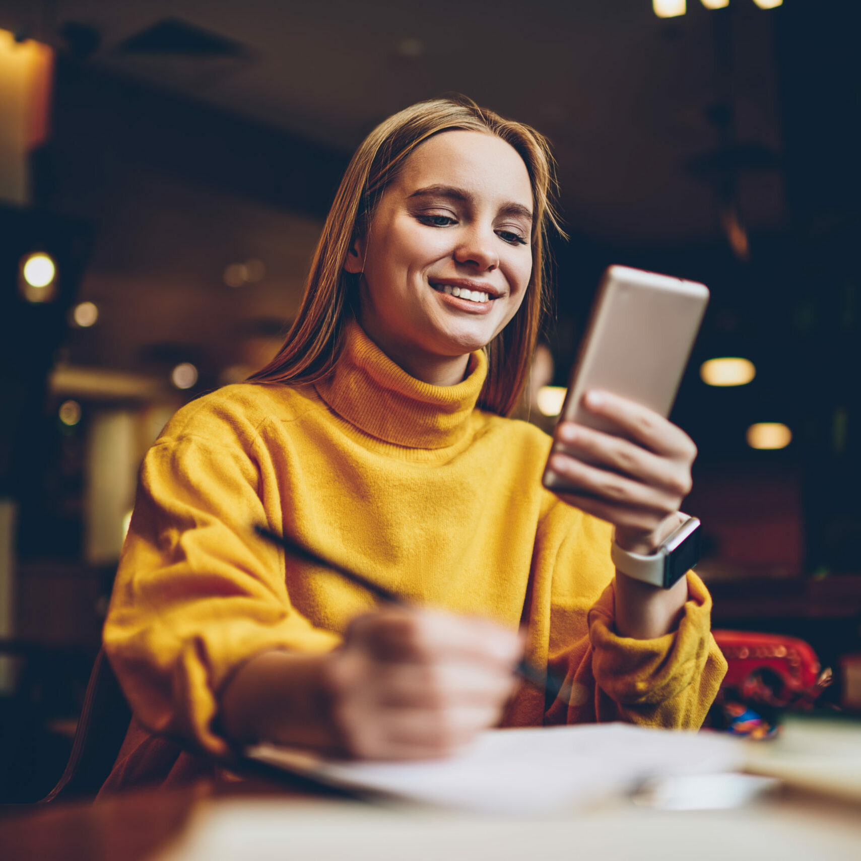 Pretty prosperous blonde woman dressed in cool yellow sweater using smartphone application for planning working schedule.Cute successful female blogger chatting with followers in social network