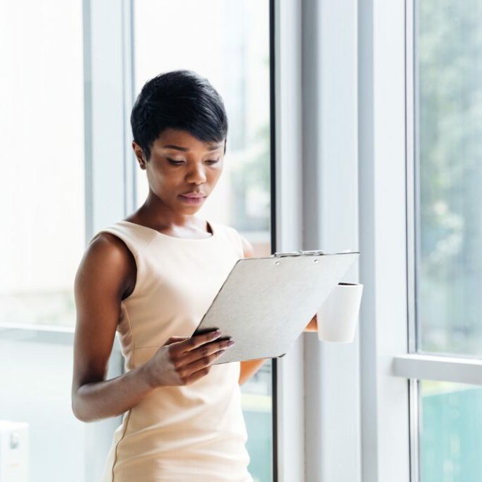 Beautiful african american young businesswoman with cup of coffee holding clipboard and reading in office